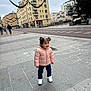 toddler, child, pink_jacket, white_sneakers, sidewalk, urban, buildings, street, holiday_decorations, pigtails, smiling, outdoor, cold_weather, pavement, people_in_background, cityscape, casual_clothing, daytime, happy, standing
