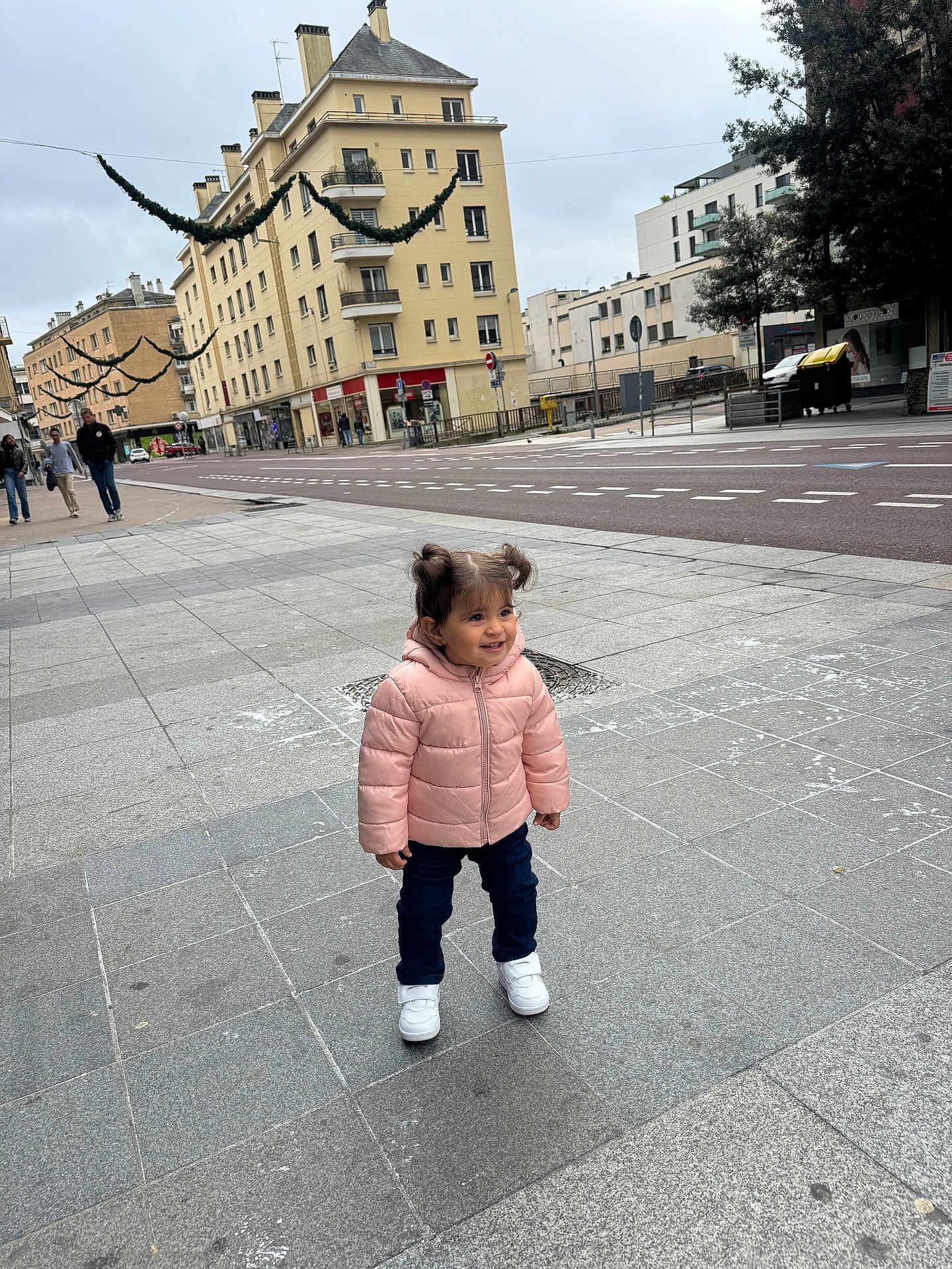 Layan a rejoint le concours — aidez-le/la à gagner de superbes lots ! toddler, child, pink_jacket, white_sneakers, sidewalk, urban, buildings, street, holiday_decorations, pigtails, smiling, outdoor, cold_weather, pavement, people_in_background, cityscape, casual_clothing, daytime, happy, standing