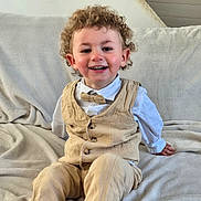 Pierre-Antoine participe au concours pour gagner de l'argent avec cette photo : beige_clothing, blanket, bowtie, casual, child, couch, curly_hair, face, furniture, happy, home, indoor, pants, person, portrait, sitting, smiling, toddler, vest, white_shirt