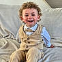 beige_clothing, blanket, bowtie, casual, child, couch, curly_hair, face, furniture, happy, home, indoor, pants, person, portrait, sitting, smiling, toddler, vest, white_shirt