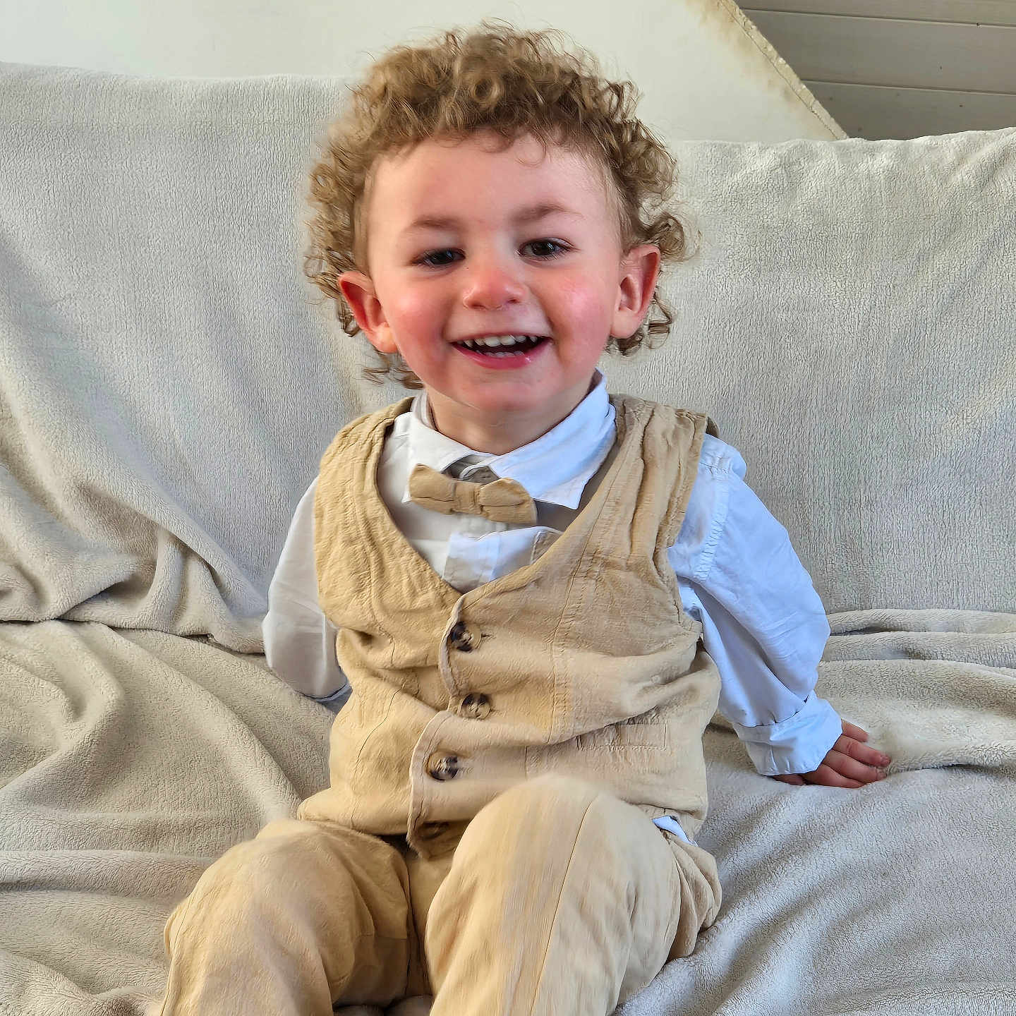 Pierre-Antoine participe au concours pour gagner de l'argent avec cette photo : beige_clothing, blanket, bowtie, casual, child, couch, curly_hair, face, furniture, happy, home, indoor, pants, person, portrait, sitting, smiling, toddler, vest, white_shirt