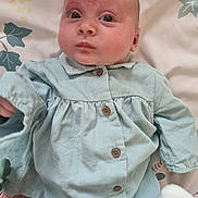 Rose is registered to the contest to win money with this photo: baby, infant, child, face, blue_clothing, buttoned_shirt, blanket, floral_pattern, lying_down, curious_expression, skin, head, eyes, mouth, hand, soft_texture, indoors, portrait, person, newborn