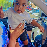Nyaire is registered to the contest to win money with this photo: adult_hand, baby, black_shoes, black_shorts, blue_flower_skirt, car_interior, child, daylight, expression, eyes, face, hand, holding, person, seat, steering_wheel, toddler, vehicle, white_shirt, window