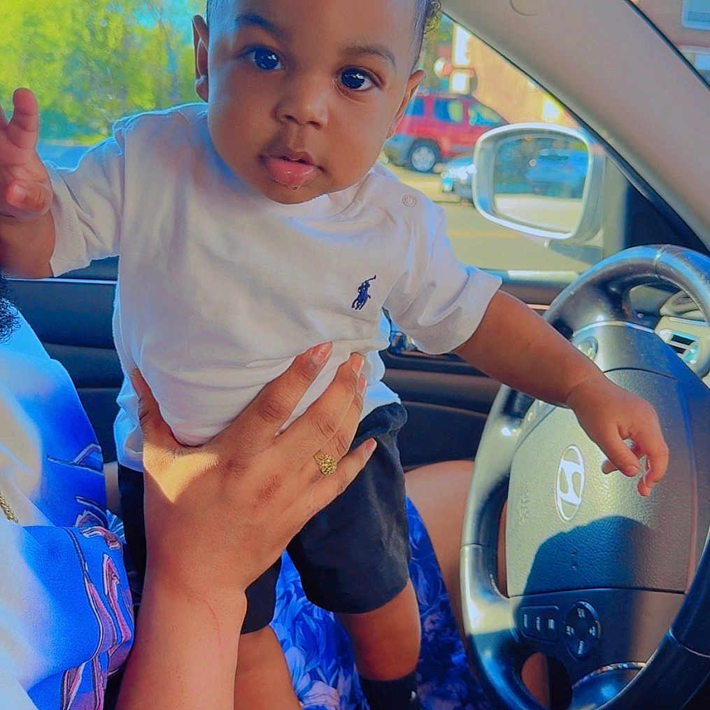 Nyaire is registered to the contest to win money with this photo: adult_hand, baby, black_shoes, black_shorts, blue_flower_skirt, car_interior, child, daylight, expression, eyes, face, hand, holding, person, seat, steering_wheel, toddler, vehicle, white_shirt, window