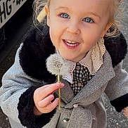 Lou participe au concours pour gagner de l'argent avec cette photo : child, toddler, girl, smiling, dandelion, coat, outdoor, street, happy, cute, hair_buns, blue_eyes, holding, person, fashion, winter_clothing, vehicle, license_plate, footwear, joyful