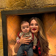 David is registered to the contest to win money with this photo: baby, woman, child, fireplace, stone_wall, smile, red_lipstick, long_hair, holding, jeans, shirt, suspenders, indoor, portrait, person, happy, casual_clothing, warm_lighting, cozy, family