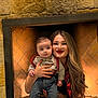 baby, woman, child, fireplace, stone_wall, smile, red_lipstick, long_hair, holding, jeans, shirt, suspenders, indoor, portrait, person, happy, casual_clothing, warm_lighting, cozy, family