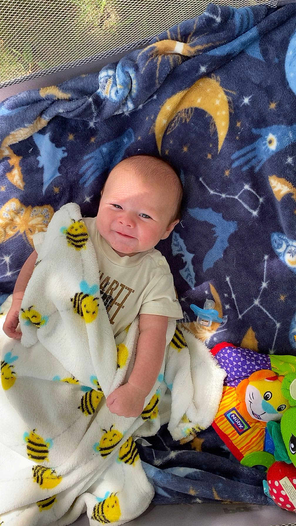 Bentley is registered to the contest to win money with this photo: baby, blanket, bee_pattern, blue_blanket, stars, moon, toys, plush_toy, lion_toy, frog_toy, pacifier, crib, mesh, infant, smiling, clothing, indoor, cute, child, playing