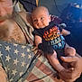 Coyote is registered to the contest to win money with this photo: baby, man, adult, holding, onesie, text_on_clothing, tattoo, tank_top, american_flag, indoor, furniture, hand, face, person, curious, casual_clothing, room, sitting, blur, closeup