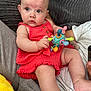 Aria joined the competition — help win amazing prizes! baby, child, infant, red_dress, toy, colorful, cushion, couch, drool, curious, person, skin, face, hand, legs, sitting, indoor, adult_arm, comfort, soft