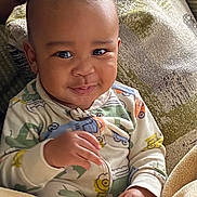 Omari joined the competition — help win amazing prizes! baby, child, smile, face, eyes, hand, onesie, clothing, blanket, couch, fabric, texture, person, infant, indoor, cozy, cute, portrait, young, happy