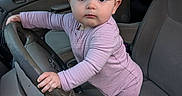 Rosalia joined the competition — help win amazing prizes! baby, child, car, steering_wheel, seat, hand, clothing, onesie, indoors, person, wide_eyes, looking_at_camera, foot, tattoo, dashboard, window, greenery, background, curious, cute