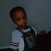 Tyron is registered to the contest to win money with this photo: toddler, child, denim_overalls, striped_shirt, necklace, cross, indoor, portrait, serious_expression, standing, young_child, person, fashion, clothing, dark_background, floor, wall, casual_wear, jewelry, cute