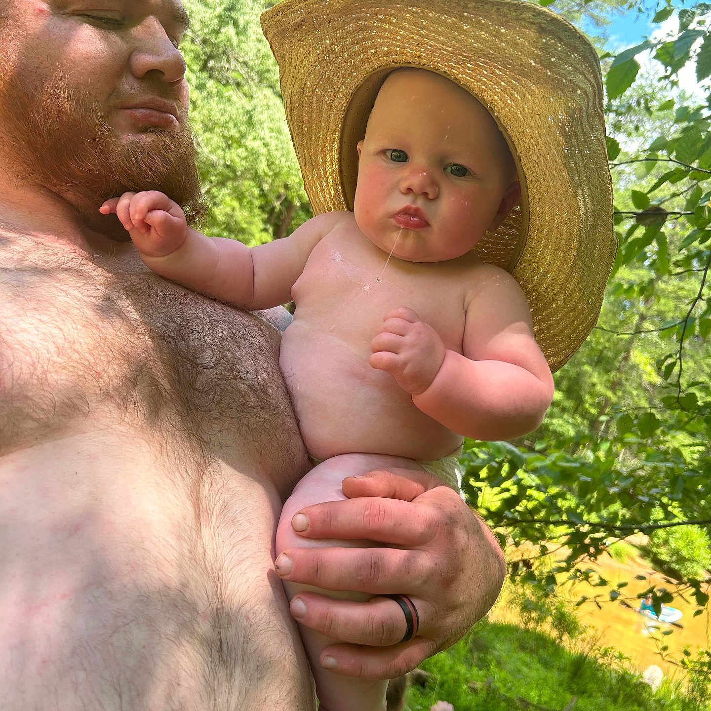 John is registered to the contest to win money with this photo: adult, baby, beard, child, daytime, drool, forest, greenery, hand, hat, holding, man, nature, outdoor, person, river, shirtless, skin, summer, sunlight