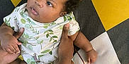 Railey is registered to the contest to win money with this photo: baby, child, infant, person, hands, hair_bow, clothing, patterned_outfit, floor_mat, checkered_pattern, yellow, black, white, green, curly_hair, lying_down, looking_sideways, skin, cute, indoor