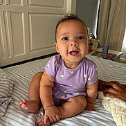 Reighlynn joined the competition — help win amazing prizes! baby, infant, child, purple_clothing, onesie, smiling, happy, sitting, bed, striped_bedspread, hand, blanket, indoor, face, cute, person, room, curtains, furniture, cozy