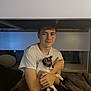 Neko joined the competition — help win amazing prizes! young_man, cat, indoor, bed, couch, window, smiling, short_sleeve_shirt, dark_hair, pet, animal, holding, seated, furniture, relaxed, home, casual, evening, resting, human