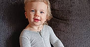 Ellia is registered to the contest to win money with this photo: barefoot, blue_eyes, casual, child, couch, cozy, curly_hair, furniture, gray_onesie, happy, holding_object, home, indoor, person, portrait, sitting, smiling, toddler, young_child, zipper