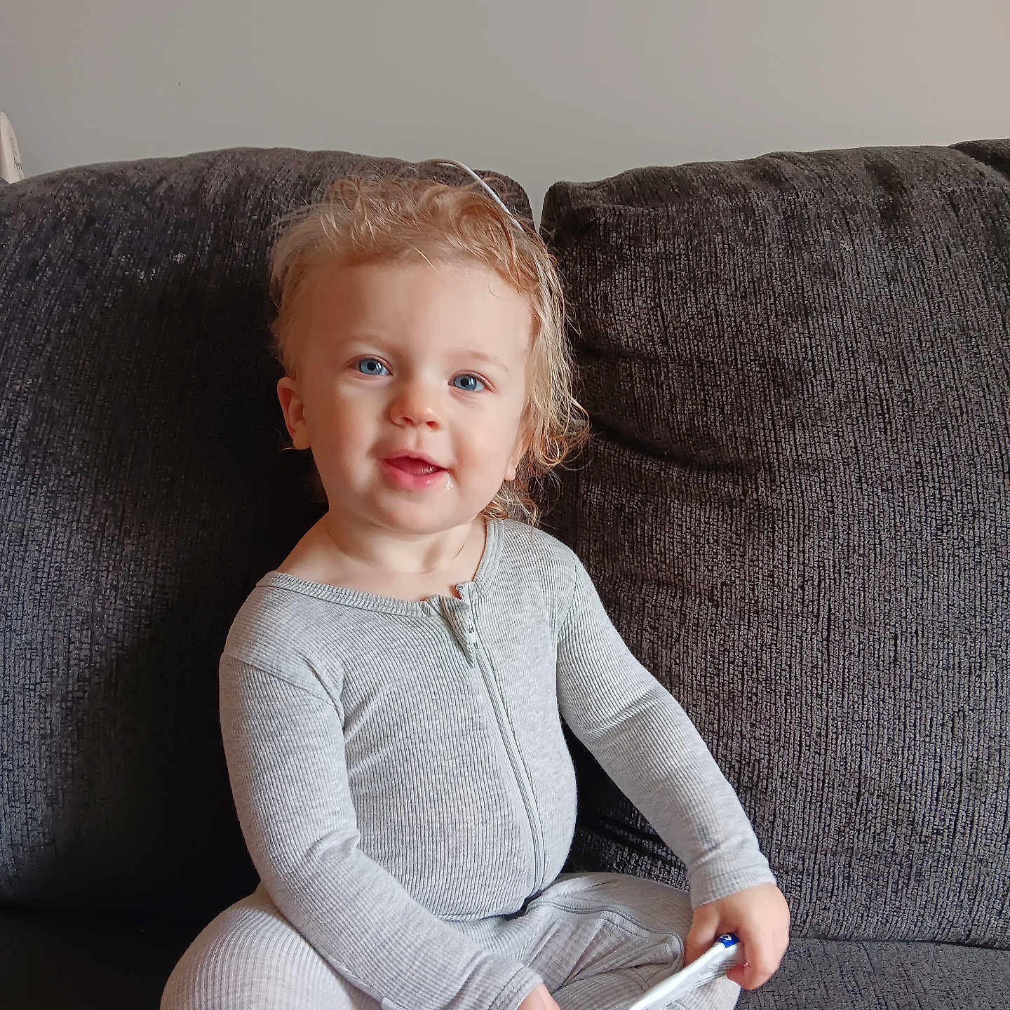 Ellia is registered to the contest to win money with this photo: toddler, child, smiling, blue_eyes, curly_hair, gray_onesie, zipper, sitting, couch, indoor, holding_object, barefoot, young_child, portrait, happy, person, furniture, cozy, casual, home