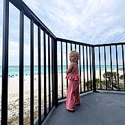 Ellia is registered to the contest to win money with this photo: child, blonde_hair, pink_clothing, balcony, railing, beach, ocean, sand, sky, cloudy, outdoor, standing, person, vacation, summer, scenic_view, coastline, nature, daytime, casual