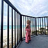 child, blonde_hair, pink_clothing, balcony, railing, beach, ocean, sand, sky, cloudy, outdoor, standing, person, vacation, summer, scenic_view, coastline, nature, daytime, casual