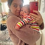 person, baby, selfie, indoor, robe, striped_clothing, holding, mirror, smartphone, home, table, chair, carpet, window, curtains, lamp, plant, ring, bracelet, intimate
