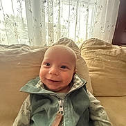 baby, child, couch, cozy, curtains, fabric, face, happy, home, indoor, infant, light, long_sleeve, portrait, seated, shadow, smiling, vest, window, zipper