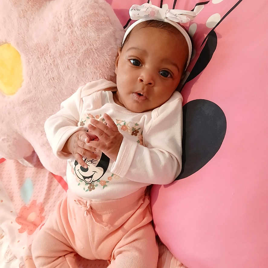 Nadiyah Kalese joined the competition — help win amazing prizes! baby, bed, blanket, bow, child, clothing, comfortable, cozy, curious, cute, face, flower_pillow, hands, headband, indoors, infant, lying_down, person, pink_pillow, soft_toys