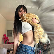 Freckles is registered to the contest to win money with this photo: black_hair, casual, cat, couch, denim_shorts, exercise_bike, furniture, holding, indoor, living_room, long_hair, orange_cat, pet, portrait, relaxed, skeleton_decoration, smile, table, white_top, woman