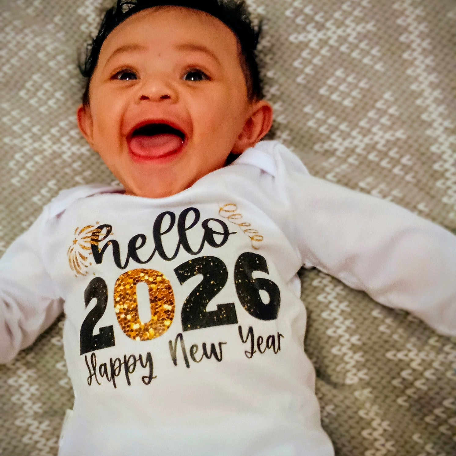 élior participe au concours pour gagner de l'argent avec cette photo : 2026, baby, blanket, celebration, child, cute, eyes, face, hair, happy, infant, laughing, lying_down, new_year, onesie, open_mouth, pattern, portrait, smiling, text_on_clothing