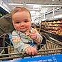 baby, child, shopping_cart, grocery_store, cute, onesie, pineapple_pattern, face, hand, smile, indoor, aisle, food_shelves, seat_belt, person, clothing, infant, curious, holding, shopping