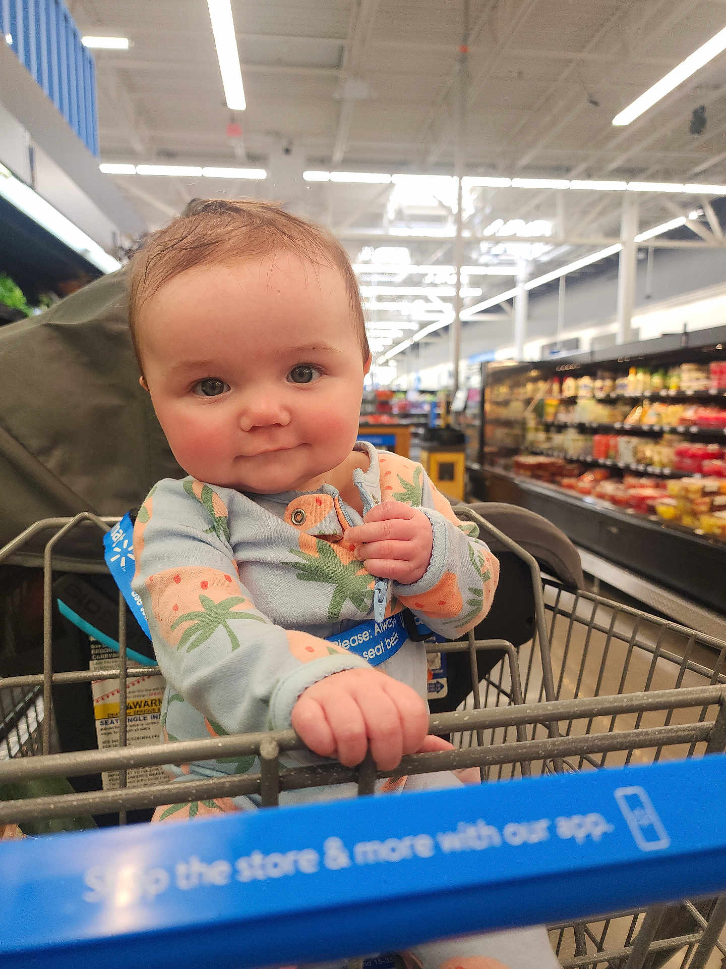Zoey is registered to the contest to win money with this photo: baby, child, shopping_cart, grocery_store, cute, onesie, pineapple_pattern, face, hand, smile, indoor, aisle, food_shelves, seat_belt, person, clothing, infant, curious, holding, shopping