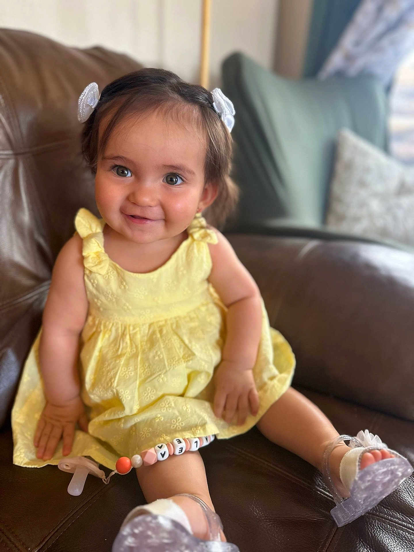 Azalea is registered to the contest to win money with this photo: toddler, child, smiling, yellow_dress, sitting, leather_couch, indoor, pacifier, hair_bows, transparent_sandals, happy, cute, portrait, baby, person, footwear, accessory, bright, comfortable, skin