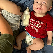 Shon is registered to the contest to win money with this photo: baby, smiling, red_shirt, diaper, bottle, blanket, blue, person, arm, child, lying_down, happy, infant, casual, indoor, cute, relaxed, skin, clothing, human