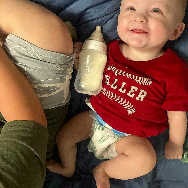Shon is registered to the contest to win money with this photo: arm, baby, blanket, blue, bottle, casual, child, clothing, cute, diaper, happy, human, indoor, infant, lying_down, person, red_shirt, relaxed, skin, smiling