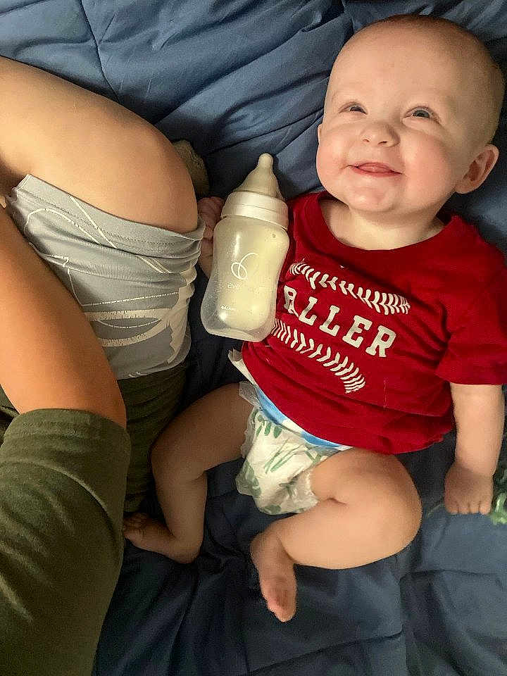 Shon is registered to the contest to win money with this photo: baby, smiling, red_shirt, diaper, bottle, blanket, blue, person, arm, child, lying_down, happy, infant, casual, indoor, cute, relaxed, skin, clothing, human