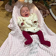 Daisy is registered to the contest to win money with this photo: baby, blanket, christmas_tree, floor, wooden_floor, toy, plush_toy, holiday, festive_clothing, socks, smiling, infant, face, person, indoors, decorations, flooring, cozy, cute, child