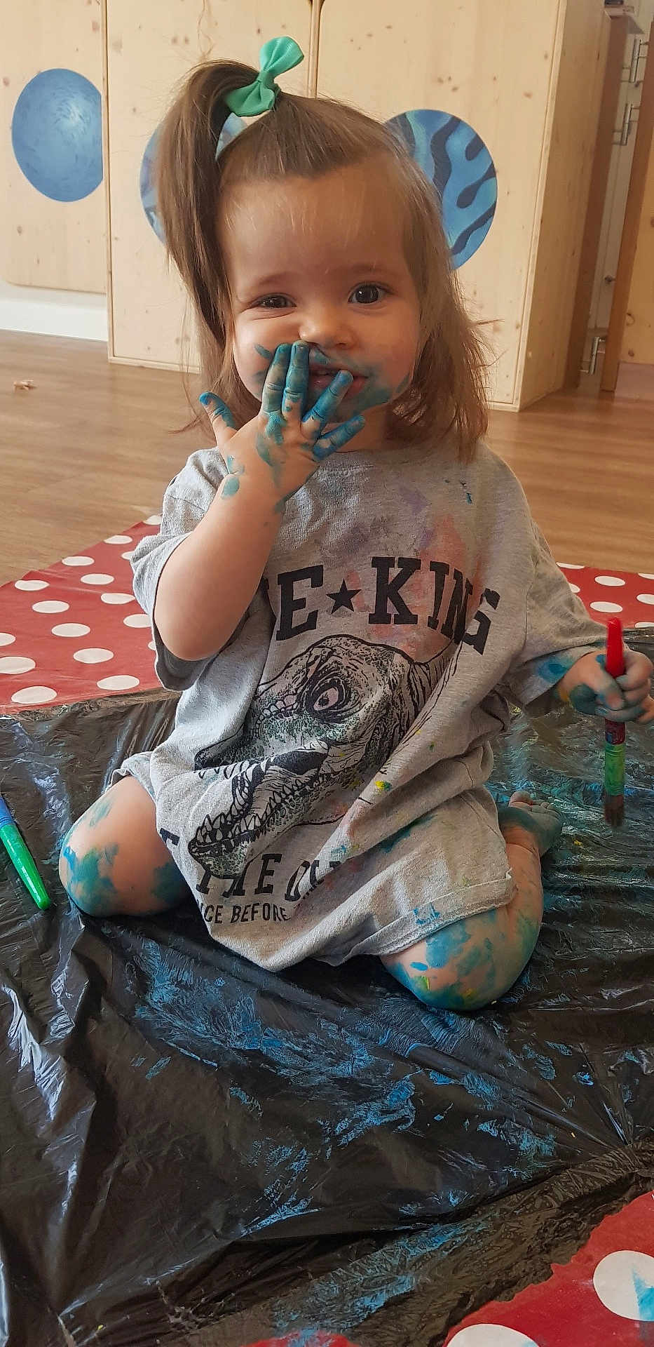 Emma a rejoint le concours — aidez-le/la à gagner de superbes lots ! toddler, child, paint, blue_paint, messy, smiling, indoor, plastic_sheet, paintbrush, clothing, hair_bow, floor, play, creative, face_paint, legs, sitting, expression, fun, art