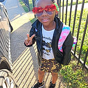 Sky is registered to the contest to win money with this photo: child, girl, sunglasses, heart_shaped, peace_sign, leather_jacket, camouflage_skirt, backpack, sidewalk, car, fence, shadow, grass, sunlight, street, casual_clothing, outdoor, smiling, fashion, portrait