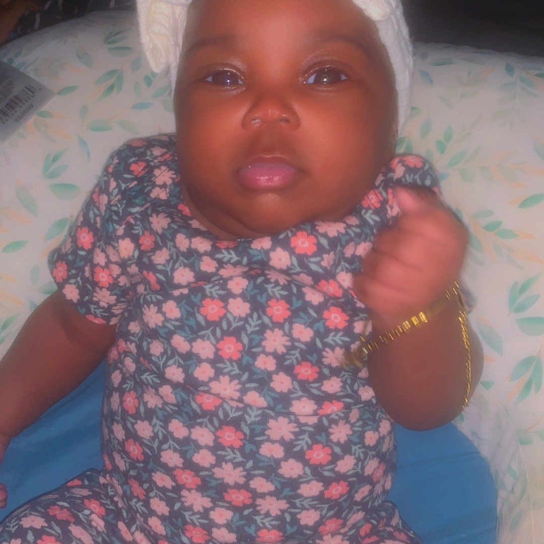 Skylar joined the competition — help win amazing prizes! adorable, baby, bracelet, child, closeup, clothing, cushion, cute, expression, fist, floral_onesie, headband, home, indoor, infant, patterned, person, portrait, sitting, soft_lighting