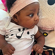 Vanity Marie is registered to the contest to win money with this photo: baby, infant, child, headband, curly_hair, white_shirt, animal_print, plush_toy, cushion, cute, indoors, portrait, looking_sideways, small_hands, seated, black_hair, pink, soft_texture, clothing, expression
