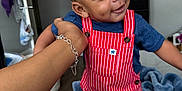 Cayden is registered to the contest to win money with this photo: toddler, child, smiling, overalls, red_clothing, striped, indoor, hand, bracelet, bed, blanket, blue_clothing, person, home, floor, doorway, cabinet, happy, cute, portrait