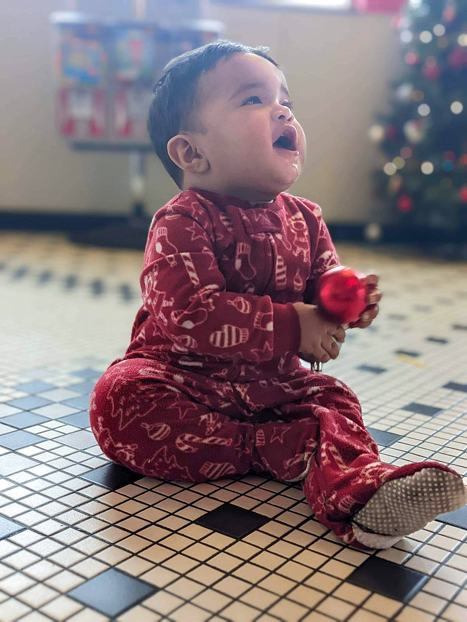 David is registered to the contest to win money with this photo: arm, baby, baby_toddler_clothing, christmas_tree, face, facial_expression, floor, flooring, hair, hairstyle, happy, head, human_body, person, photograph, purple, skin, sleeve, smile, standing