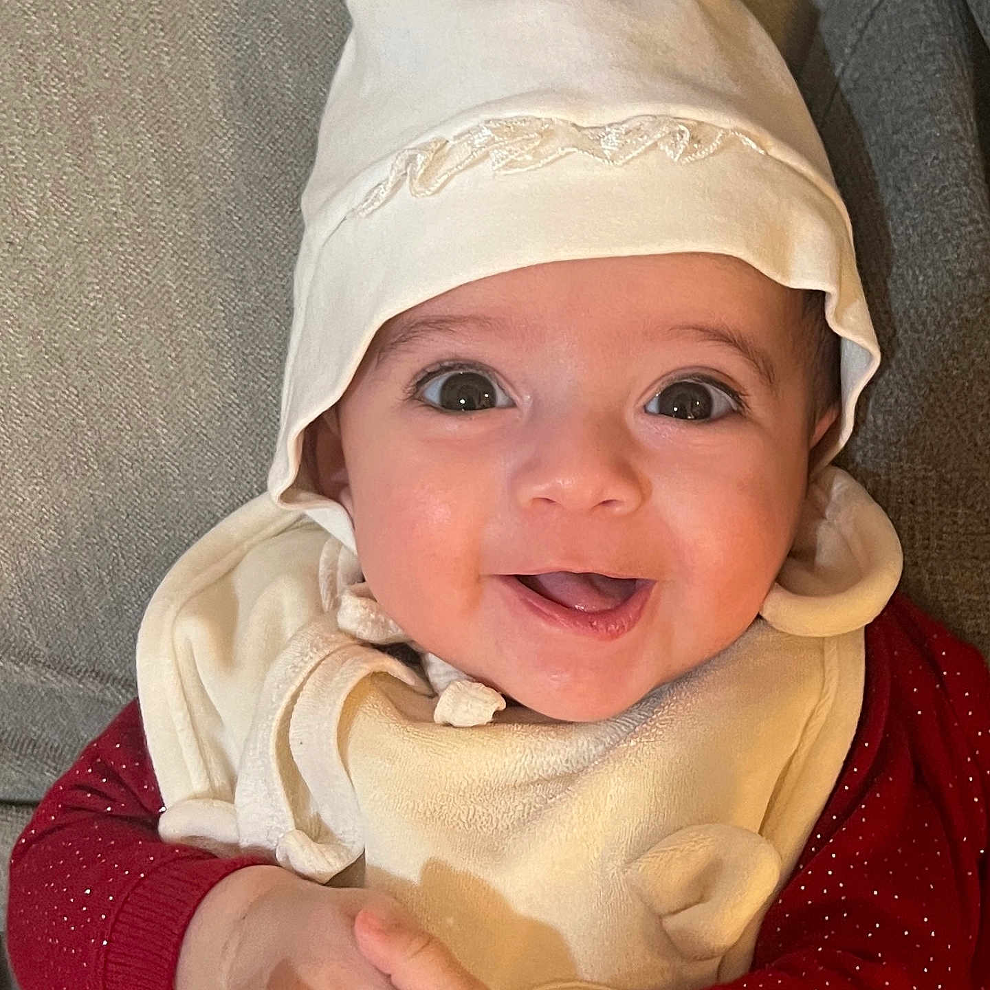 Dhalia joined the competition — help win amazing prizes! baby, bib, big_eyes, bonnet, cheek, child, closeup, clothing, couch, fabric, hands, happy, hat, indoor, infant, mouth, portrait, red_sweater, skin, smile
