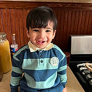 Ghasan is registered to the contest to win money with this photo: child, toddler, boy, smile, portrait, indoor, kitchen, wooden_paneling, countertop, stove, jar, bottle, striped_shirt, polo_collar, buttons, dark_hair, sitting, happy, closeup, eyes