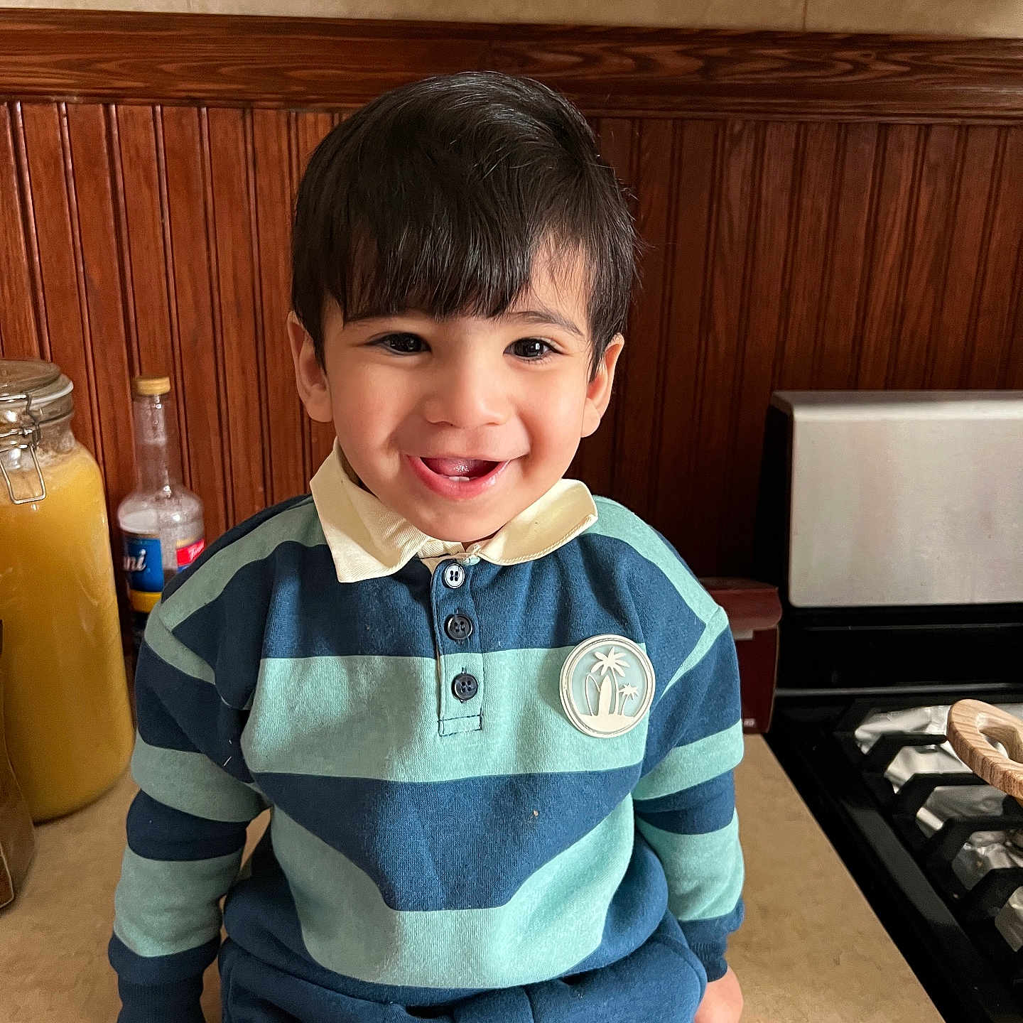 Ghasan is registered to the contest to win money with this photo: bottle, boy, buttons, child, closeup, countertop, dark_hair, eyes, happy, indoor, jar, kitchen, polo_collar, portrait, sitting, smile, stove, striped_shirt, toddler, wooden_paneling