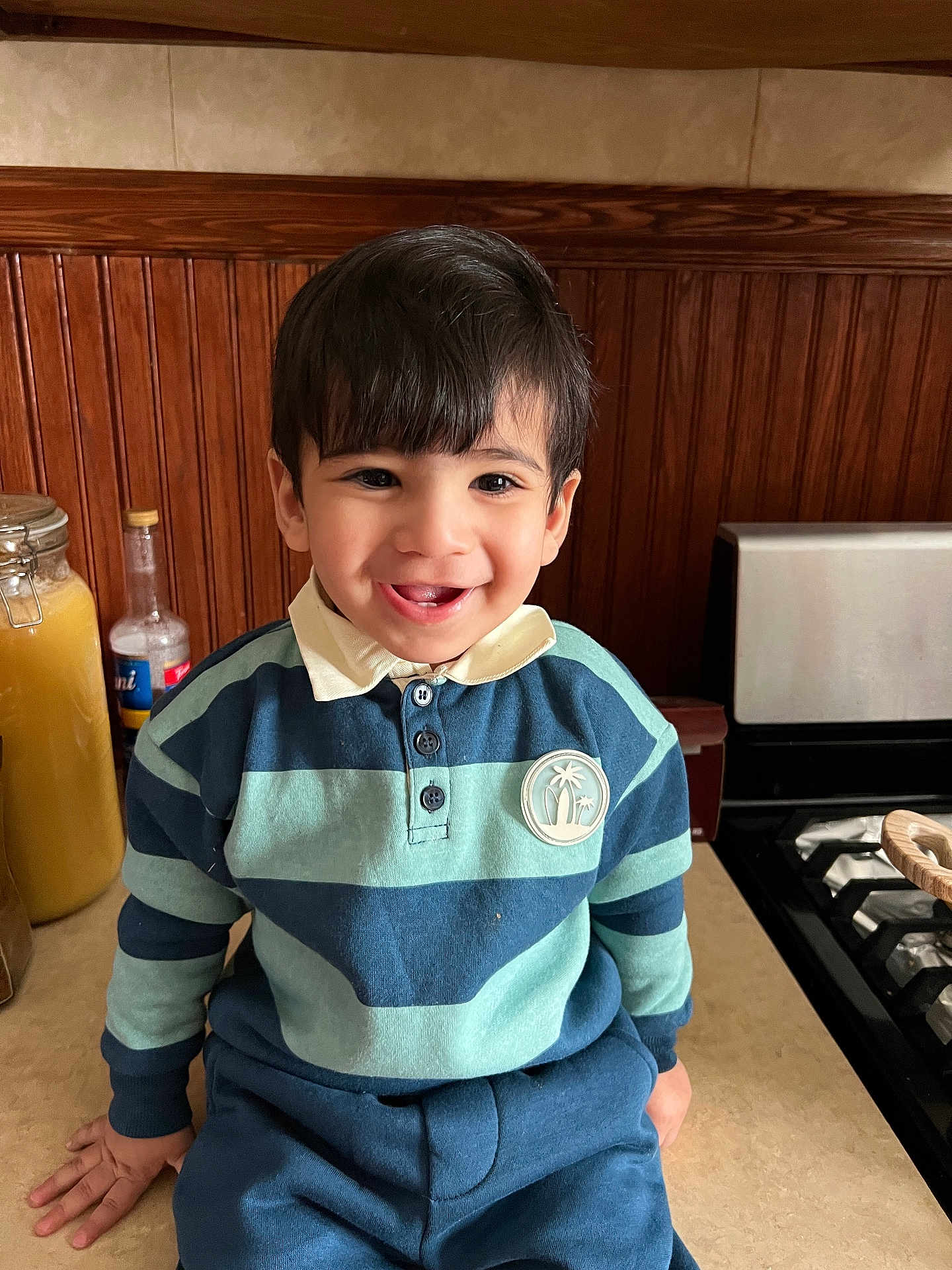 Ghasan is registered to the contest to win money with this photo: child, toddler, boy, smile, portrait, indoor, kitchen, wooden_paneling, countertop, stove, jar, bottle, striped_shirt, polo_collar, buttons, dark_hair, sitting, happy, closeup, eyes