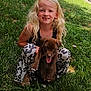 child, girl, puppy, dog, grass, outdoor, smiling, happy, blonde_hair, curly_hair, black_top, patterned_pants, tongue_out, pet, playing, nature, sunlight, brick_wall, crouching, cute