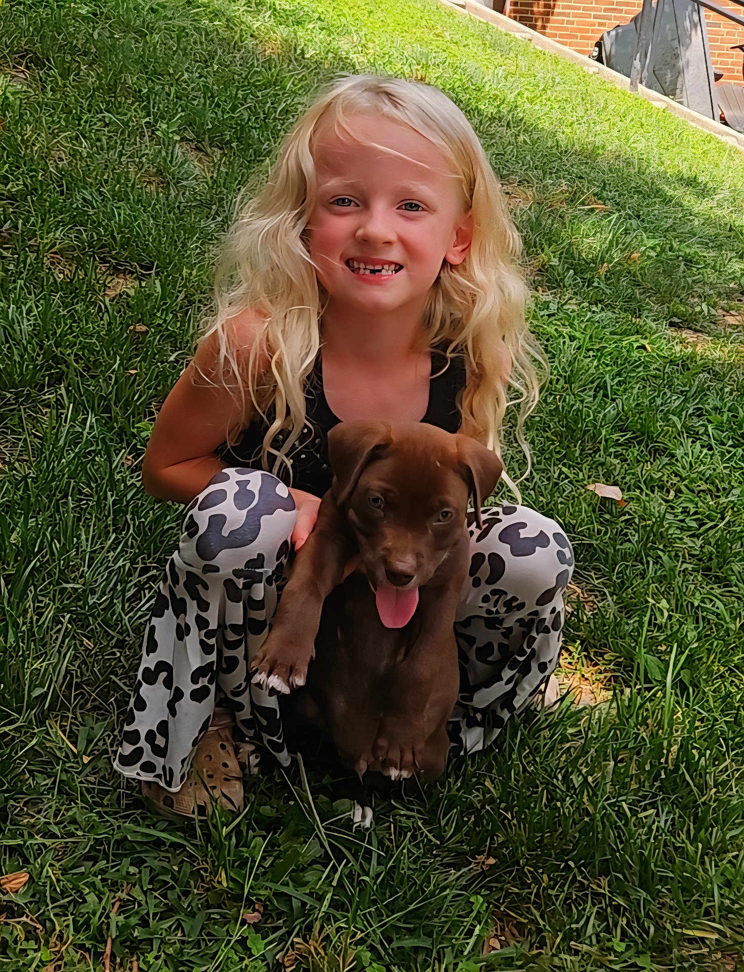 Dollar joined the competition — help win amazing prizes! child, girl, puppy, dog, grass, outdoor, smiling, happy, blonde_hair, curly_hair, black_top, patterned_pants, tongue_out, pet, playing, nature, sunlight, brick_wall, crouching, cute