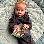 Freddy is registered to the contest to win money with this photo: baby, infant, child, milestone_card, 5_months_old, plush_mat, soft_texture, red_hair, cozy_clothing, striped_sock, cute, portrait, indoor, holding_object, looking_at_camera, baby_pose, monkey_illustration, milestone, young_child, comfortable